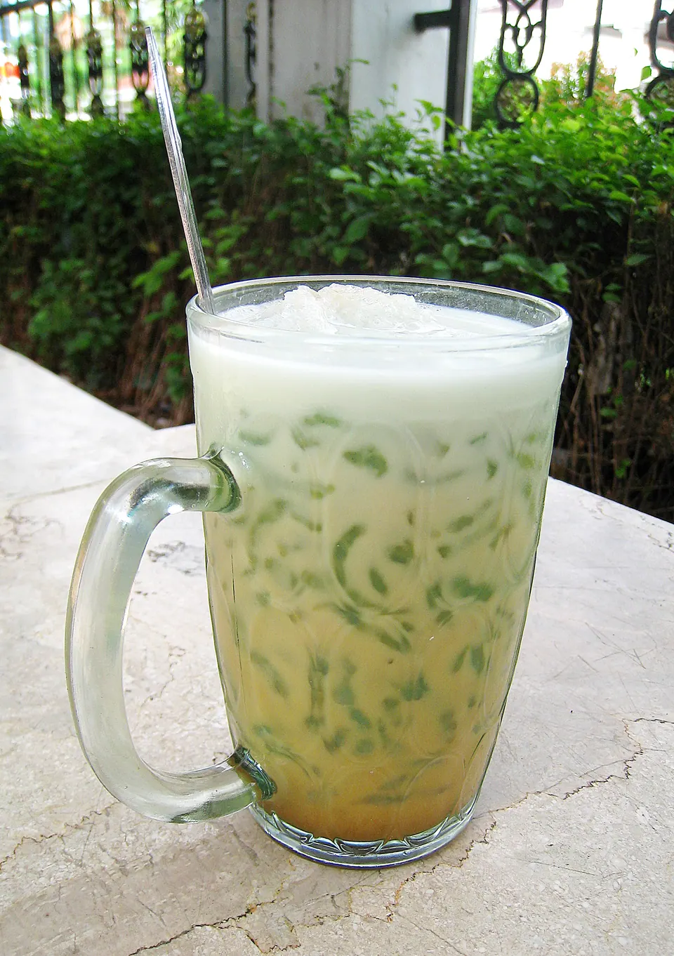 Segelas es cendol dengan untaian hijau dalam santan, difoto di luar ruangan dengan langit dan dedaunan